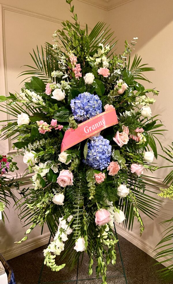 Express your sympathy with a standing easel spray featuring blue hydrangeas, pink roses, and lush greenery from Flowerland Florist in Sulphur Springs, TX. A stunning floral tribute for funerals and memorial services. Local delivery available.